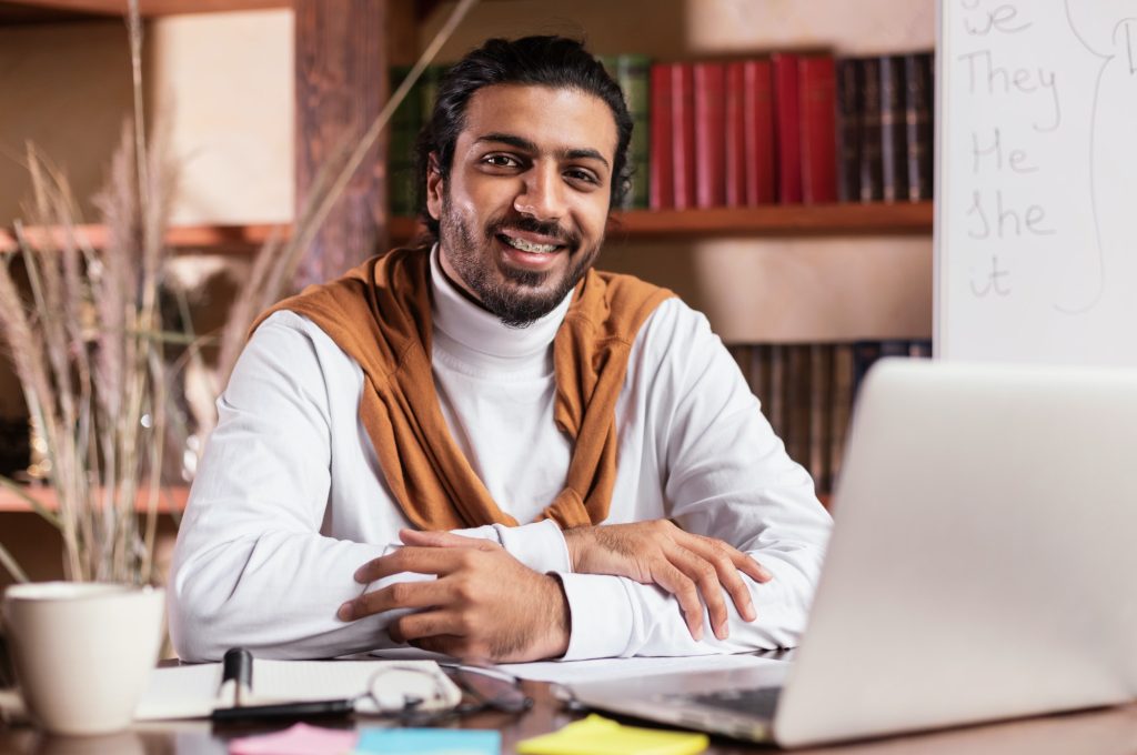 successful-indian-teacher-sitting-at-laptop-posing-in-library-indoors.jpg